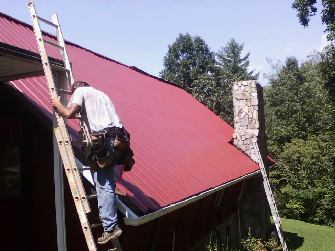 Roofer on a ladder beside a red metal roof and stone chimney — Soffit & Fascia Repair by Roofing Brothers in Cape Girardeau